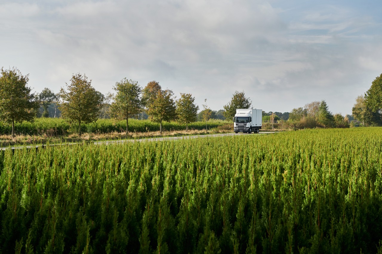 Bild på en vit Scania-lastbil som kör på en väg med en åker i framkant
