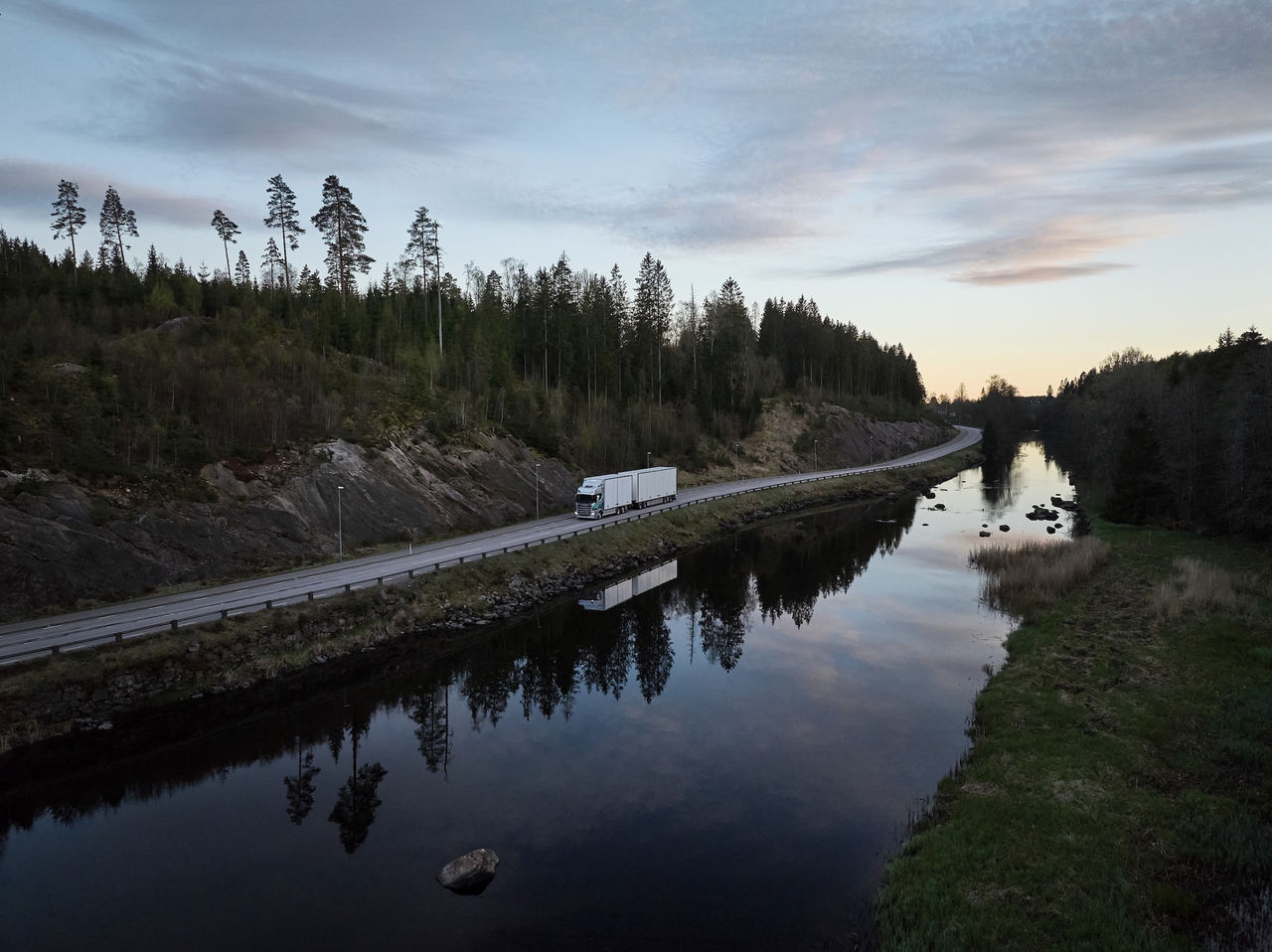 Aerial view of a Scania truck driving along a narrow road surrounded by river and forest on sunset