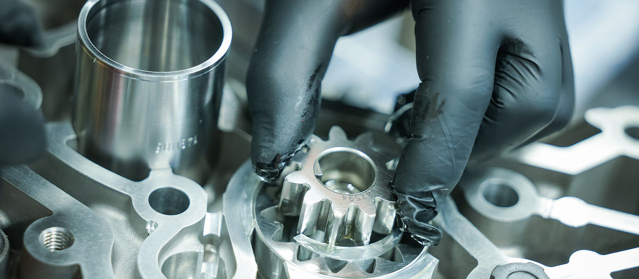 A hand wearing a black protective glove places a gear into a machined metal component.